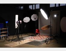 Fotostudio mit dunklem Hintergrund, mehreren Stativen mit Studiobeleuchtung, einem Kamerastativ, einem roten Stuhl und einem bunten Teppich auf dem Boden. Links steht ein kleiner Wagen mit Monitoren und Kabeln.