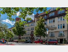 Wohngebäude im Stil des frühen 20. Jahrhunderts mit detailreichen Fassaden, Satteldächern und mehreren Fenstern, umgeben von Bäumen an einer gepflasterten Straße mit parkenden Autos; ein Straßenschild mit der Aufschrift „Südenstraße“ ist im Vordergrund sichtbar.