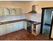 Eine einfache Küche mit Holzschränken in hellgrünem Farbton, einer braunen Arbeitsplatte und einem eingebauten Gasherd mit Dunstabzugshaube. Der Boden besteht aus dunklem Holzlaminat, und die Wände sind teils mit weißen Fliesen verkleidet. Eine offene Tür führt zu einem angrenzenden Raum mit einem Fenster und Heizkörper.