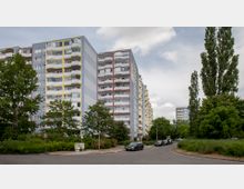 Mehrstöckige Wohngebäude mit grauer Fassade und bunten Balkonen (gelb und rot) in einer Wohnsiedlung, umgeben von Bäumen, Grünflächen und parkenden Autos entlang einer Straße. Der Himmel ist bewölkt.