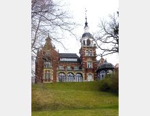 Historisches Gebäude mit roter Backsteinfassade und gelben Sandsteinverzierungen, einem markanten Turm mit Kuppel und mehreren Rundbogenfenstern. Das Gebäude steht auf einem leicht ansteigenden, grasbewachsenen Hügel und ist von Bäumen umgeben.