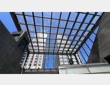 Blick durch ein modernes Glasdach mit Gitterstruktur auf mehrere Hochhäuser und den blauen Himmel. Im Vordergrund befinden sich Gebäudeteile mit Glas- und Betonfassaden.