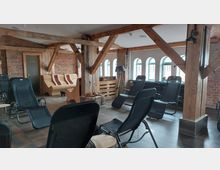 Ein gemütlicher Ruheraum mit Holzträgern, Backsteinwänden und großen, bogenförmigen Fenstern, ausgestattet mit schwarzen Liegestühlen und einer Holzbank mit Kissen. Im Raum steht eine Stehlampe neben einem Korb mit Handtüchern.