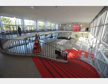 Großer, lichtdurchfluteter Veranstaltungsraum mit einer geschwungenen Treppe, die von einem oberen Rundgang in den unteren Bereich führt. Der Raum hat große Fensterfronten, weiße Sitzreihen vor einer Bühne und eine zentrale Kunstinstallation in Form einer beleuchteten Skulptur.