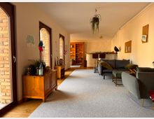 Langer Wohnraum mit großen, bogenförmigen Fenstern auf der linken Seite und Holzmöbeln entlang der Wand. Der Raum ist mit Teppichboden ausgelegt, umfasst ein graues Sofa, Sessel, einen kleinen Tisch und eine Theke mit Barhockern im Hintergrund. Eine Hängepflanze und Wanddekorationen ergänzen die Einrichtung.