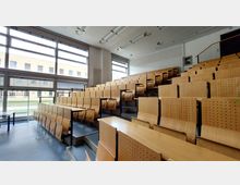 Ein moderner Hörsaal mit ansteigenden Sitzreihen aus perforierten Holzstühlen und integrierten Klapptischen. Große Fenster an der linken Wand lassen viel Tageslicht herein und bieten Ausblick auf ein Gebäude mit gelblicher Klinkerfassade.