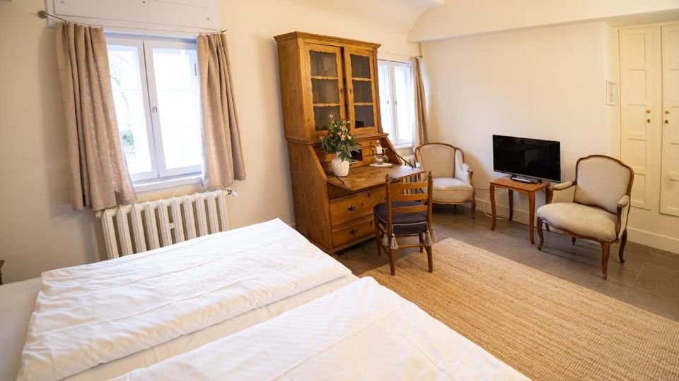 Helles Gästezimmer mit zwei Betten, großen Fenstern mit Vorhängen und Heizkörper darunter. Ein Holzsekretär mit Vitrine, zwei Polstersessel, ein kleiner Tisch mit Fernseher sowie ein heller Teppich auf Fliesenboden ergänzen die Einrichtung.