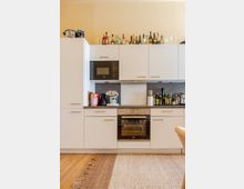 Moderne Einbauküche mit weißen Schrankfronten, dunkler Arbeitsplatte und grauer Rückwand, ausgestattet mit Mikrowelle, Dunstabzug und Backofen. Auf der Arbeitsfläche und auf den Oberschränken stehen mehrere Flaschen, Küchenutensilien und Gläser; davor liegt ein Teppich auf Holzboden.