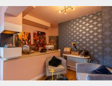 Wohnzimmer mit dekorativer Einrichtung, rosa Wänden und blauer Tapete mit Schuppenmuster. Im Raum befinden sich graue Sessel, ein runder Couchtisch, Wandregale, Bilder auf einer Ablage und ein stilvolles Grammophon auf einem Beistelltisch.