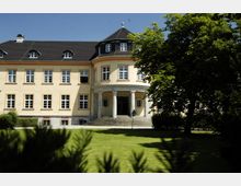 Helle historische Villa mit cremefarbener Fassade, zahlreichen Fenstern und einem zentralen Haupteingang mit Säulen und einer kleinen Treppe, umgeben von einem gepflegten Rasen und schattenspendenden Bäumen.
