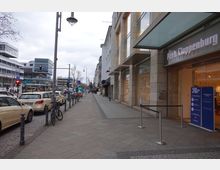 Straßenszene in einer Einkaufsgegend mit Bürgersteig, parkenden Autos und einer Filiale von Peek & Cloppenburg mit gläsernem Eingangsbereich auf der rechten Seite. Im Hintergrund sind mehrere moderne Gebäude, darunter das Schloss-Straßen-Center, und Straßenlaternen zu sehen.