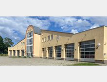 Modernes Feuerwehrgebäude mit beigefarbener Fassade, mehreren großen Toren für Einsatzfahrzeuge und einem zentralen Eingangsbereich mit hoher Glasfront und gewölbtem Dach. Das Gebäude steht auf einer gepflasterten Fläche und ist von vereinzeltem Grün umgeben.