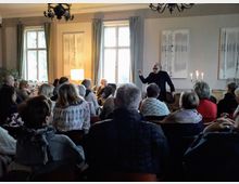 Eine Gruppe von Menschen sitzt in einem stilvoll eingerichteten Raum mit hohen Fenstern und Vorhängen, während ein Mann vorne steht und spricht. Im Hintergrund sind ein Kronleuchter, Gemälde an der Wand und Kerzen auf einem Tisch zu sehen.
