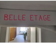 Ein Innenraum mit einem grauen Durchgang, über dem in pinker Schrift "BELLE ETAGE" steht. Im Hintergrund befinden sich helle Räume mit hölzernen Trennwänden und einer großen Pflanze.