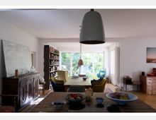 Wohnbereich mit modernen und klassischen Elementen, darunter ein Holztisch mit verschiedenen Stühlen, ein gläserner Buffetschrank und Bücherregale. Im Hintergrund befinden sich große Fenster, die einen Blick auf einen grünen Garten ermöglichen, während Schalen mit Obst im Vordergrund auf einer Theke stehen.