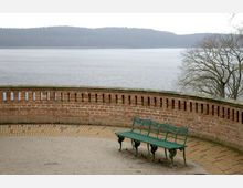 Eine geschwungene Backsteinmauer mit kleinen rechteckigen Öffnungen begrenzt eine erhöhte Aussichtsplattform, auf der eine grüne Metallbank steht. Im Hintergrund ist ein ruhiger See mit einer bewaldeten Uferlinie zu sehen, und rechts ragt ein kahler Baum ins Bild.