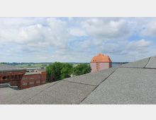 Blick über mehrere Dächer auf eine ländliche Landschaft mit Feldern, Bäumen und einem Backsteingebäude mit rotem Ziegeldach im Hintergrund. Am Horizont sind ein Fluss und bewölkter Himmel erkennbar.