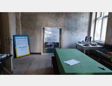 Ein einfacher Raum mit teilweise abblätternder Wandfarbe, in dem ein Tisch mit grünem Tuch und ein Zettel darauf stehen. Im Hintergrund stehen Plakate und Farbdosen auf einem weiteren Tisch, während rechts große Fenster viel Tageslicht hereinlassen.