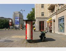 Straßenszene mit einem kleinen Platz, umgeben von Gebäuden mit heller Steinfassade. Im Vordergrund eine Litfaßsäule und ein geparktes Motorrad, links im Bild Bäume, eine Ampel, und weitere Gebäude, darunter ein Bau mit Gerüst und Werbung.