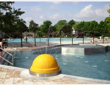 Außenbereich eines Schwimmbades mit mehreren Becken, einem gelben Wasserspiel in der Vordergrundmitte und klar strukturierten Flächen. Im Hintergrund Bäume, eine Steinbrücke und Personen, die sich im und um das Wasser aufhalten.