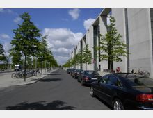Straße mit parkenden Autos entlang einer modernen Gebäudezeile aus grauem Beton. Rechts davon stehen vereinzelt Bäume, Fahrräder und Fußgänger, während der Himmel teilweise bewölkt ist.