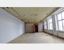 Ein leerer, renovierungsbedürftiger Raum mit hohen Decken, einem ornamentierten Stuckelement in der Mitte der Decke, abblätternden Wänden sowie mehreren großen, rechteckigen Fenstern auf der rechten Seite, die natürliches Licht hereinlassen.