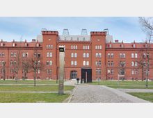 Backsteingebäude mit historischer Fassade im neugotischen Stil, gekennzeichnet durch Türme, Rundfenster und Zinnen; davor ein gepflasterter Weg, Rasenflächen, Bäume ohne Laub und eine moderne Stele als Skulptur.