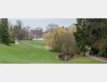 Parklandschaft mit weitläufigen Rasenflächen, einem Teich mit kleiner Holzbrücke und mehreren Bäumen, darunter eine Trauerweide. Im Hintergrund ist ein Gebäude im historischen Stil zu sehen, rechts führt ein Spazierweg entlang einer Baumreihe.