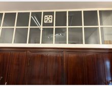 Innenansicht eines Raums mit dunkler Holzvertäfelung und einer darüberliegenden Glasfensterreihe in weißem Rahmen, die in einen angrenzenden Bereich blickt. Hinter den Scheiben sind Deckenleuchten und ein an der Decke montiertes Gerät sichtbar.