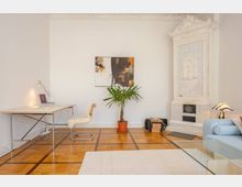 Helles Wohnzimmer mit einem Schreibtisch und einem Stuhl mit Fellauflage, einem abstrakten Bild an der weißen Wand, einem kleinen Palmengewächs in einem Topf sowie einem weißen, reich verzierten Kachelofen. Der Boden besteht aus Holzparkett, und ein Teil eines hellblauen Sofas sowie eines Glastisches ist sichtbar.