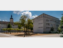 Platz mit trockenem Rasen, einzelnen Bäumen und gepflasterten Wegen, umgeben von einem modernen Gebäude mit rechteckigen Fenstern auf der rechten Seite und einer älteren Gebäudereihe mit einem Kirchturm im Hintergrund. Der Himmel ist überwiegend blau mit einigen Wolken.