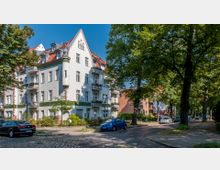 Wohnhaus im Stil der Gründerzeit mit weißer Fassade, roten Ziegeldächern und kleinen Balkonen, umgeben von Bäumen und einer gepflasterten Straße mit parkenden Autos. Die grüne Straßenallee vermittelt ein ruhiges, städtisches Wohngebiet.