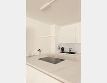 Moderne Küche mit minimalistischer Gestaltung. Eine helle Kücheninsel mit integrierter Induktionskochplatte steht im Vordergrund, während im Hintergrund weiße Schränke, ein schwarzes Spülbecken und ein maritimer Wasserhahn zu sehen sind.