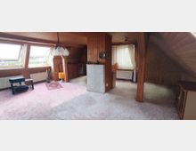 Wohnzimmer in einem Dachgeschoss mit holzvertäfelten Wänden und Decken. Der Raum verfügt über schräge Wände mit Dachfenstern, einen Teppichboden, eine Stehlampe, Möbel und ein Fenster mit weißem Vorhang.