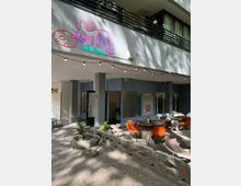 Außenbereich eines Cafés mit runden Betontischen und orangefarbenen Stühlen, umgeben von dekorativen Betonbarrieren mit roten Akzenten. An der Fassade darüber sind eine bunte Katzenillustration und kleine Lichterketten befestigt; die Umgebung ist schattig mit Bäumen.