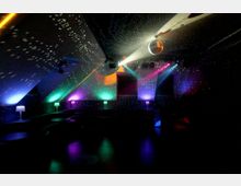 Innenraum eines Nachtclubs mit schräger Decke und einer Discokugel, die Lichtpunkte über Wände und Decke streut. Bunte Scheinwerfer und indirekte Beleuchtung erhellen eine dunkle Tanzfläche mit niedrigen Sitzbereichen und kleinen Tischlampen entlang der Seiten.