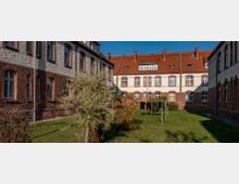 Innenhof eines historischen Gebäudekomplexes mit roten Ziegelfassaden und hellem Putz, eingefasst von drei Gebäudeflügeln. Im Hof befinden sich eine gepflegte Rasenfläche, einzelne Bäume und ein Wäscheständer mit aufgehängter Kleidung.