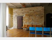 Ein Raum mit einer Wand aus unverputzten Ziegelsteinen, einer schweren Metalltür, mehreren orangefarbenen Polstersitzen und blauen Stühlen. Im Hintergrund sind eine offene Dusche und helle Vorhänge sichtbar.