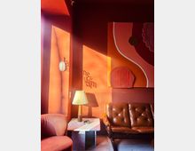 Warm-toned lounge corner with deep coral walls, a brown tufted leather sofa, a pink upholstered chair, and a small marble-topped side table with a pleated lamp. Abstract textured wall art and a round wall sconce are lit by strong sunlight casting long shadows across the wall.