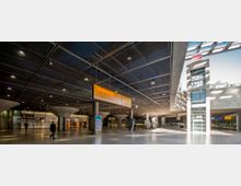 Geräumige unterirdische Bahnhofs- oder Passagierhalle mit moderner Architektur, gekennzeichnet durch eine Gitterdecke, Betonpfeiler, Informationsschilder und gläserne Aufzüge; im Hintergrund befinden sich Geschäfte und Zugänge zu Bahnsteigen.