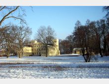 Schloss in einer winterlichen Parklandschaft mit schneebedecktem Boden und kahlen Bäumen. Das Gebäude ist hellgelb, mehrstöckig und verfügt über einen mittelalterlich anmutenden Turm.