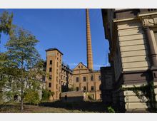 Industriekomplex mit historischen Backsteingebäuden, darunter ein hoher Schornstein, umgeben von Bäumen und überwucherter Vegetation. Rechts im Bild ist ein neoklassizistisches Gebäude mit verzierten Fassadenelementen zu sehen.