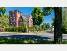 Backsteingebäude mit großen Rundbogenfenstern und einem Satteldach, umgeben von einer Hecke und einem Metallzaun; das Gebäude befindet sich an einer Kreuzung in einer grünen, von Bäumen gesäumten Straßenlandschaft.