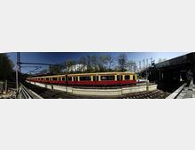 Eine Panoramaansicht eines S-Bahnhofs mit einem gelb-roten Zug auf den Gleisen. Im Hintergrund sind Bäume und ein Gebäude zu sehen, rechts führt eine Treppe zu einer Fußgängerbrücke, und links befinden sich Bahninfrastruktur und ein Halteverbotsschild.