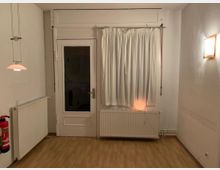 Ein kleiner Raum mit hellem Laminatboden, einer weißen Tür mit Glaseinsatz und einem bodenlangen Vorhang. Links befindet sich ein Heizkörper mit einem darüber hängenden Lampenschirm, rechts ein weiterer Heizkörper unter der Fensterbank, daneben eine leuchtende Wandlampe. Ein Feuerlöscher ist an der linken Wand montiert.