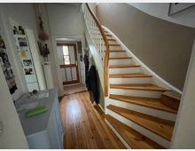 Flur im Eingangsbereich eines Hauses mit Holzfußboden und einer geschwungenen Holztreppe mit weißem Geländer, die nach oben führt. Am Ende des Flurs steht eine Haustür mit Glaseinsatz; seitlich sind eine Kommode und Wandhaken für Jacken zu sehen.