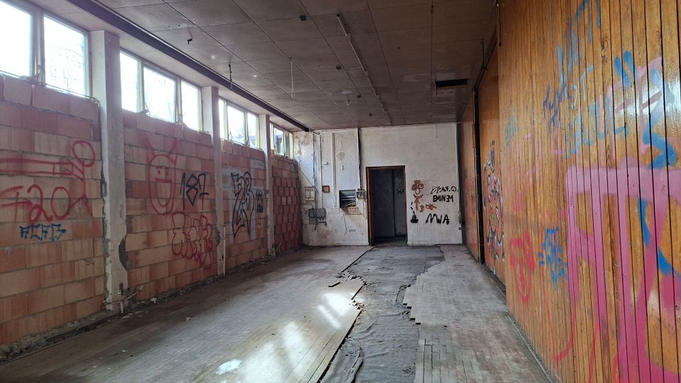 Langer, leerer Innenraum einer verlassenen Industriehalle mit hohen Fensterreihen links, rauen Ziegelwänden und einer holzverkleideten Wand rechts, die mit Graffiti besprüht ist. Der Boden ist teilweise beschädigt und aufgerissen, am Ende des Raums befindet sich eine offene Tür in einer weißen Wand.
