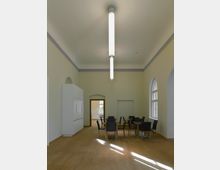 Ein heller Raum mit Holzfußboden und pastellgelben Wänden, ausgestattet mit mehreren Holztischen und schwarzen Stühlen. Große Rundbogenfenster lassen Tageslicht ein, und eine lange, schmale Deckenleuchte beleuchtet den Raum.