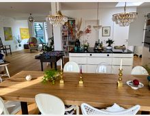 Offener Wohnbereich mit hellen Möbeln, Holzboden und einer modernen Küche mit weißer Arbeitsinsel. Im Vordergrund steht ein Holz-Esstisch mit goldenen Kerzenständern, im Hintergrund sind Bücherregale, ein Kicker und eine Sitzecke mit Dekorationen sichtbar.