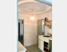 Kleine Küchenzeile in einem hellen Raum mit weiß gestrichenen Wänden und grauem Boden. Links eine Tür zum Schlafzimmer, rechts ein Spülbecken mit weißem Unterschrank und darüber offene Regale mit Geschirr. Eine zentral hängende Lampe wirft kreisförmige Lichtmuster an die Decke.